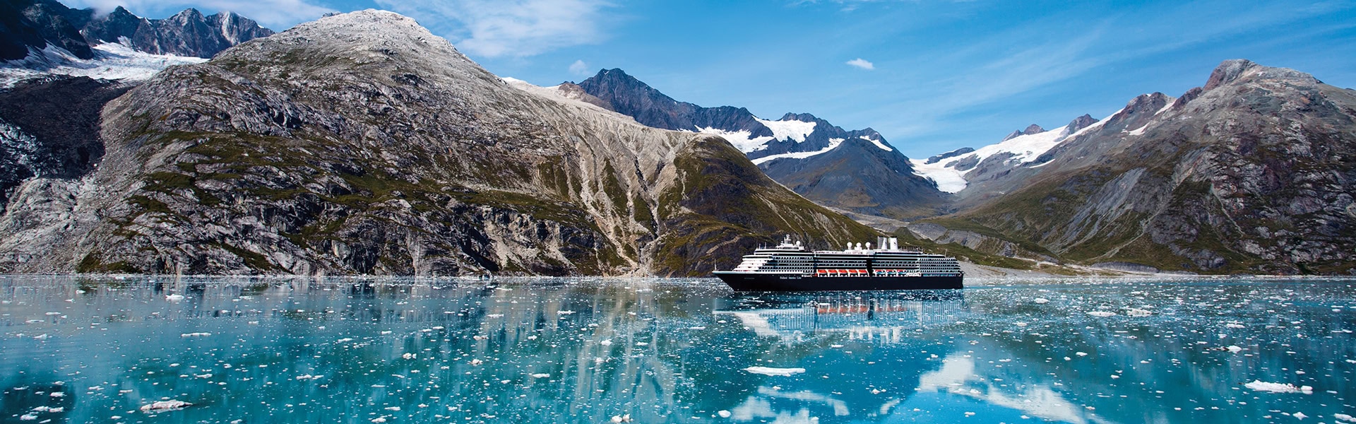 Ein großes Kreuzfahrtschiff der Holland America Line fährt durch ein ruhiges, türkisfarbenes Fjordgewässer in Alaska, das mit zahlreichen kleinen Eisschollen und Eisbergen übersät ist. Im Hintergrund erheben sich monumentale, karge Felswände mit schneebedeckten Gipfeln und sichtbaren Gletschern unter einem klaren, blauen Himmel. Das Schiff und die Berge spiegeln sich deutlich auf der Wasseroberfläche.