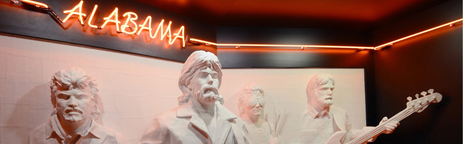 Vier weiße, detailreiche Skulpturen der Bandmitglieder der Country-Gruppe "Alabama" sind als Halbreliefs vor einer hellen Wand dargestellt. Die Figuren zeigen die Musiker mit ihren charakteristischen Frisuren und Bärten der 1980er Jahre, wobei die rechte Figur eine Bassgitarre hält. Über den Skulpturen leuchtet ein großer, orangefarbener Neon-Schriftzug mit dem Wort „ALABAMA“ in Schreibschrift. Das warme, rötliche Licht der Leuchtschrift erhellt den Raum und setzt sich als leuchtende Linie an den dunklen Wänden im Hintergrund fort.