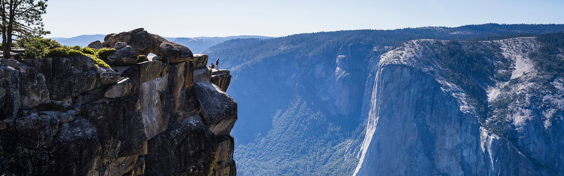 Zwei Personen stehen auf dem felsigen Vorsprung des Taft Point im Yosemite-Nationalpark und blicken in ein tiefes, bewaldetes Tal. Im Hintergrund ragt die gewaltige, senkrechte Granitwand des El Capitan unter einem klaren, blauen Himmel empor.