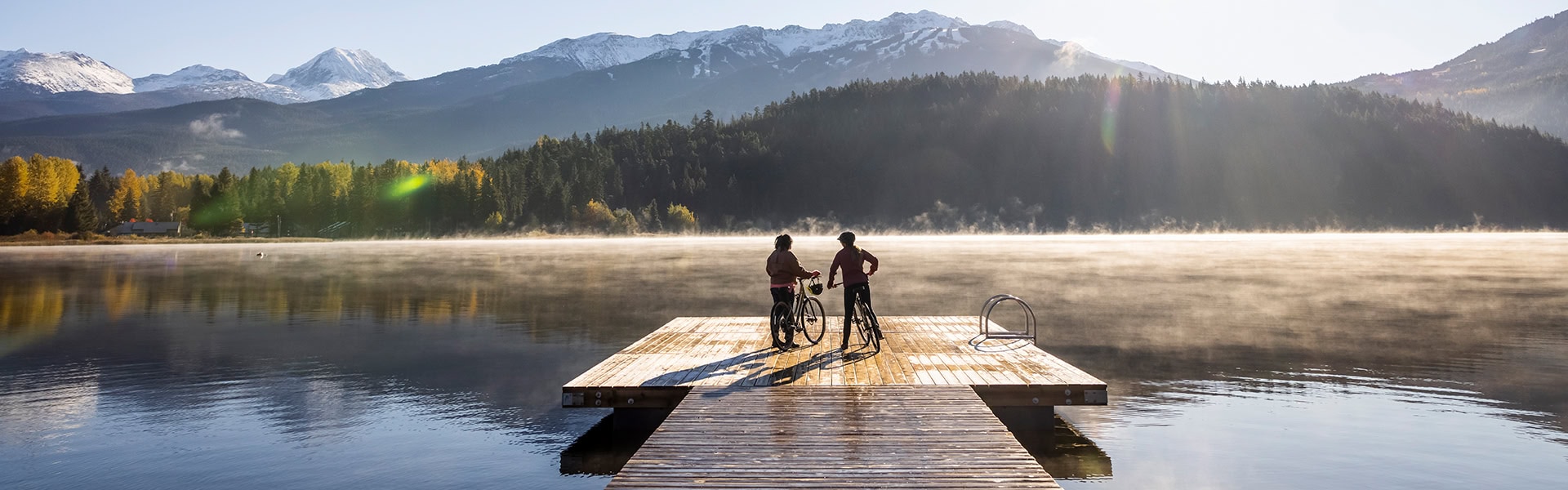 Zwei Personen stehen mit ihren Fahrrädern auf einem Holzsteg an einem ruhigen See am frühen Morgen, über dem Nebel liegt. Im Hintergrund erstrecken sich dichte Wälder und schneebedeckte Berge unter klarem Himmel.