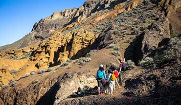 Eine Wandergruppe mit Rucksäcken zieht über einen steinigen Pfad durch eine karge, rötlich-braune Vulkanlandschaft mit steilen Felswänden unter blauem Himmel.