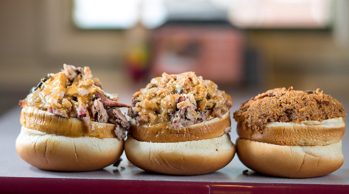 Drei verschiedene Barbecue-Sandwiches stehen nebeneinander auf einer Theke, reichlich belegt mit gezupftem und geschnittenem Fleisch sowie Sauce.