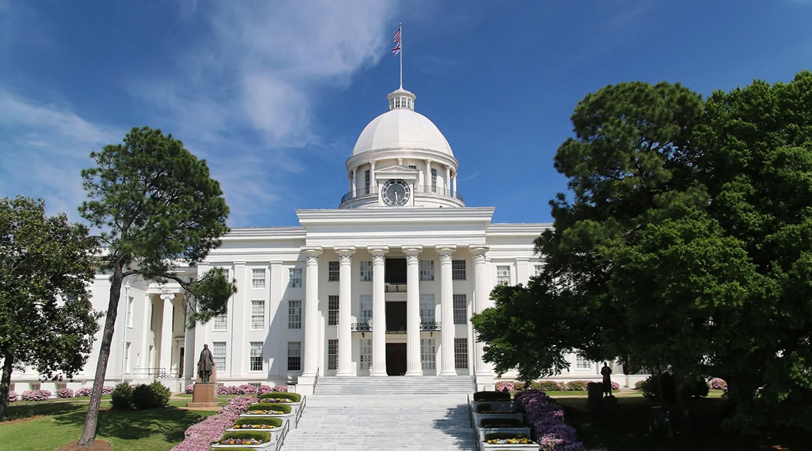 Die Vorderansicht des weißen Alabama State Capitol mit seiner markanten Kuppel, einer großen Freitreppe und korinthischen Säulen unter blauem Himmel.