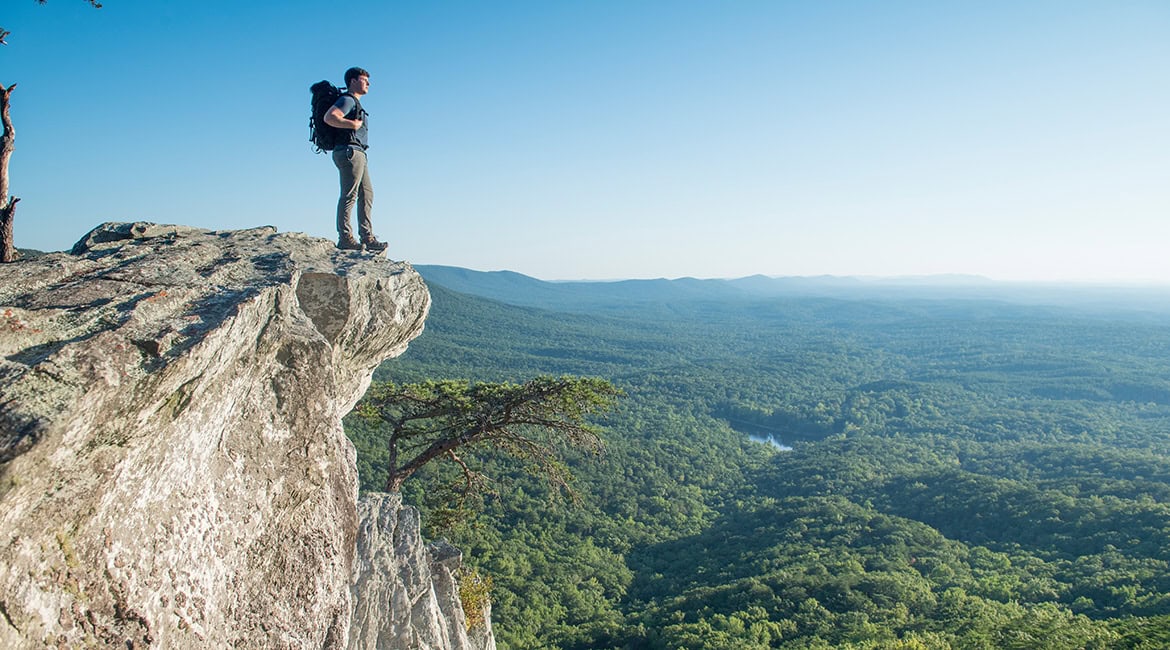 Ein Wanderer mit großem Rucksack steht auf der Kante eines steilen Felsvorsprungs und blickt über ein weites, dicht bewaldetes Tal unter klarem blauem Himmel.