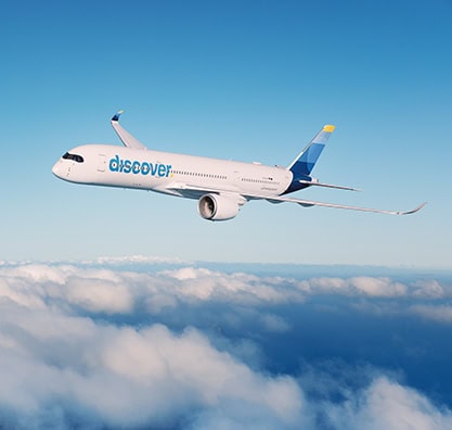 Ein weißes Passagierflugzeug der Discover Airlines fliegt bei klarem, blauem Himmel über einer dichten, weißen Wolkendecke.