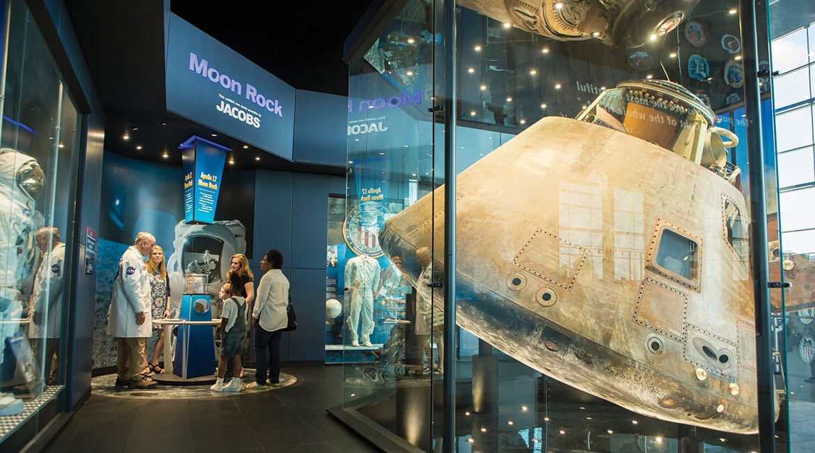 Besucher in einer Raumfahrtausstellung betrachten Mondgestein, während im Vordergrund eine originale Apollo-Kommando-Kapsel hinter einer großen Glasscheibe präsentiert wird.