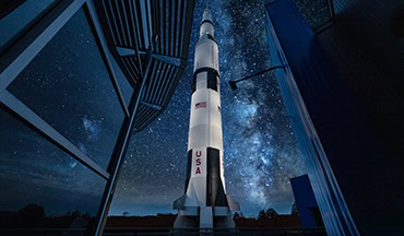 Eine beleuchtete Saturn-V-Rakete steht senkrecht im Freien zwischen modernen Museumsgebäuden unter einem sternenklaren Nachthimmel mit der Milchstraße.