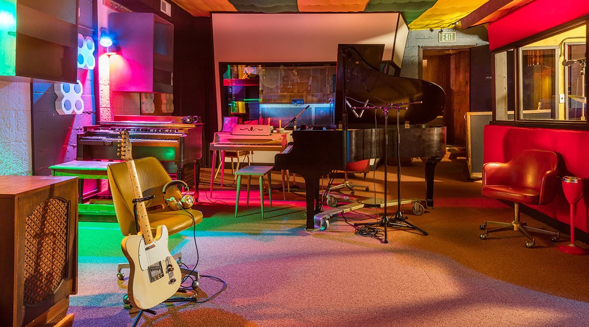 Ein bunt beleuchteter Aufnahmeraum eines Tonstudios mit verschiedenen Instrumenten wie einem Konzertflügel, Keyboards und einer E-Gitarre auf einem Ständer.
