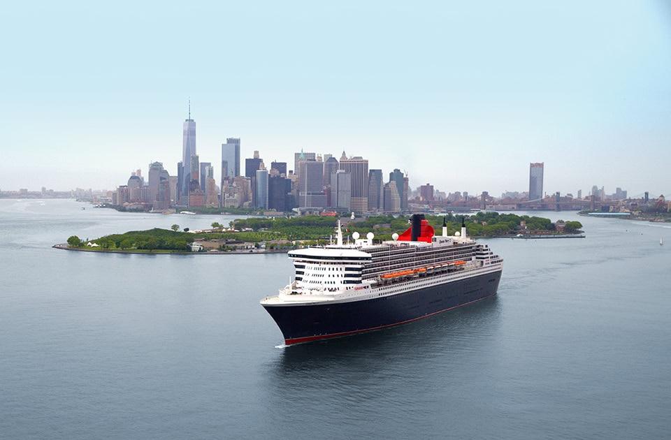 Die Queen Mary 2 fährt auf einem breiten Gewässer vor der Skyline von Lower Manhattan in New York. Das Ozeanschiff mit dunklem Rumpf, weißen Decks und rotem Schornstein steuert nach rechts, während im Hintergrund das One World Trade Center und die Wolkenkratzer der Stadt unter einem hellen, leicht bewölkten Himmel aufragen. Links im Mittelgrund ist Governors Island zu sehen.