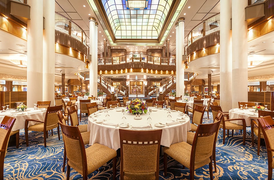 Das prunkvolle, mehrstöckige Britannia Restaurant an Bord der Queen Mary 2. Im eleganten Speisesaal stehen festlich gedeckte, runde Tische mit weißen Tischtüchern und Holzstühlen auf einem blau-gemusterten Teppich mit Wellenmotiven. Große, weiße Säulen ragen empor zu geschwungenen Balkonen der oberen Ebenen, während im Hintergrund eine geschwungene Freitreppe nach oben führt. Die Decke wird von einer riesigen, beleuchteten Glaskuppel im Art-déco-Stil dominiert.