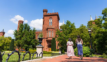 Eine sonnige Außenaufnahme des historischen, aus roten Ziegeln erbauten Smithsonian Institution Buildings (bekannt als das „Castle“) in Washington, D.C.. Im Vordergrund spazieren zwei Frauen auf einem rötlich gepflasterten Spazierweg des Enid A. Haupt Gardens in Richtung des schlossähnlichen Gebäudes mit seinen markanten Türmen. Der Weg wird von einer kunstvollen, schwarzen schmiedeeisernen Beeteinfassung gesäumt. Üppige grüne Bäume, gepflegte Hecken und ein dekoratives schwarzes Pflanzgefäß auf einem Sockel umgeben die Szenerie unter einem strahlend blauen Himmel mit vereinzelten weißen Wolken.