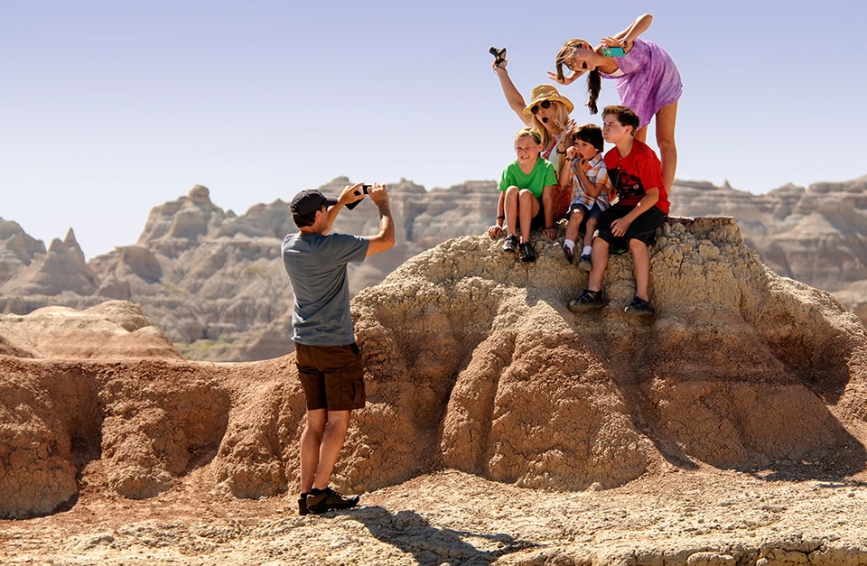 Eine Familie posiert für ein Foto in einer kargen, felsigen Wüstenlandschaft mit erodierten Felsformationen im Hintergrund. Im Vordergrund links steht ein Mann mit einer dunklen Kappe, einem grauen T-Shirt und braunen Shorts von hinten, der mit seinem Smartphone eine Gruppe fotografiert. Die Gruppe besteht aus einer Frau mit Sonnenhut und fünf Kindern, die gut gelaunt und mit teils lustigen Grimassen auf einem erhöhten, hellbraunen Erdhügel sitzen und stehen. Das Wetter ist sonnig und der Himmel klar.