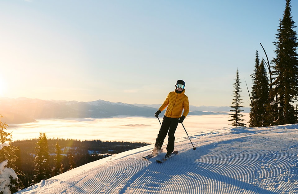 Ein Skifahrer fährt im Gegenlicht der tiefstehenden Sonne eine frisch präparierte Skipiste hinunter. Er trägt eine leuchtend gelbe Daunenjacke, eine dunkle Skihose, einen Helm und eine verspiegelte Skibrille. Im Hintergrund erstreckt sich eine weite Berglandschaft über einer dichten Wolken- oder Nebeldecke, während rechts am Pistenrand schneebedeckte Nadelbäume stehen.