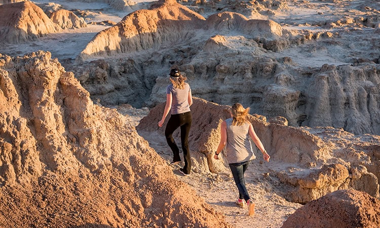 Eine Aufnahme aus leicht erhöhter Position zeigt zwei Frauen von hinten, die sich ihren Weg durch die zerklüftete, erodierte Felslandschaft des Badlands-Nationalparks bahnen. Die vordere Frau trägt ein graues T-Shirt, eine schwarze Hose und eine dunkle Kappe, während die hintere Frau in einem grauen Tanktop und Jeans geht. Die felsigen Hügel und tiefen Furchen der Badlands sind im warmen Licht der tiefstehenden Sonne zu sehen, was lange Schatten wirft und die rotbraunen und beigen Erdtöne der Formationen betont.