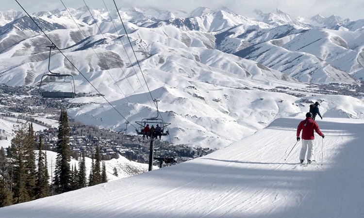 Eine Aufnahme aus erhöhter Position zeigt Skifahrer auf einer schneebedeckten Piste mit Blick auf eine Seilbahn und eine weite, verschneite Gebirgslandschaft. Im rechten Vordergrund fährt ein Skifahrer in einer roten Jacke und weißer Hose hangabwärts, während weiter hinten ein zweiter Skifahrer in dunkler Kleidung zu sehen ist. Links im Bild verläuft ein Sessellift parallel zur Piste, auf dem Personen transportiert werden; ein leerer Sessel schwebt weiter oben. Im Hintergrund erstrecken sich unzählige schneebedeckte Berggipfel unter einem leicht bewölkten Himmel, und im Tal ist eine kleine Ortschaft inmitten von Bäumen erkennbar.