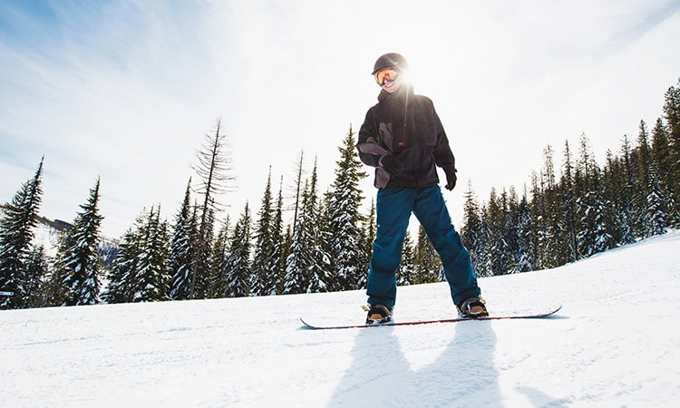 In der Mitte des Bildes ist ein Snowboarder. Hinter ihm sieht man Tannen und eine Schneelandschaft