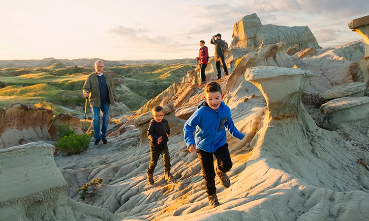 Eine Aufnahme in der warmen Abendsonne zeigt eine mehrteilige Familie beim Wandern durch eine zerklüftete Felslandschaft (Badlands) mit markanten Hoodoo-Formationen. Im Vordergrund laufen zwei kleine Jungen begeistert einen hellen, erodierten Felshang hinunter – der ältere in einer leuchtend blauen Jacke, der jüngere dahinter in einer dunklen Jacke. Weiter hinten auf dem Pfad folgt ein älterer Mann mit Brille, olivgrüner Jacke und Wanderstöcken. Auf einer höheren Felsrippe im Hintergrund stehen zwei weitere Erwachsene; einer blickt durch ein Fernglas in die Ferne. Die sanfte Hügellandschaft im Hintergrund ist von grünem Gras bewachsen und wird vom goldenen Licht der untergehenden Sonne angestrahlt.