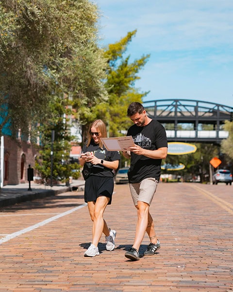 Zwei Urlauber laufen mit Karte und Smartphone über eine historische Ziegelsteinstraße in Ybor City, Tampa, mit der Centro Ybor Fußgängerbrücke im Hintergrund.