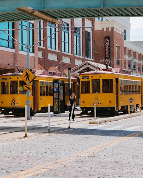 Zwei historische gelbe Straßenbahnen stehen an einer Station in Ybor City, während eine Passantin dazwischen hindurchgeht.