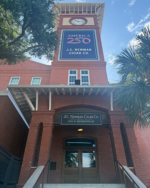 Historische Backsteinfassade der J.C. Newman Cigar Co. mit einem großen Uhrenturm und einem „America 250“-Banner unter blauem Himmel.