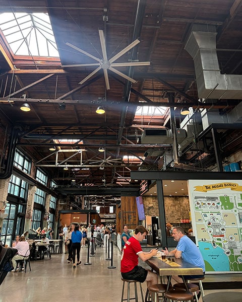 Menschen sitzen und gehen in der historischen Industriehalle der Armature Works Food Hall unter einer hölzernen Decke mit großen Ventilatoren.