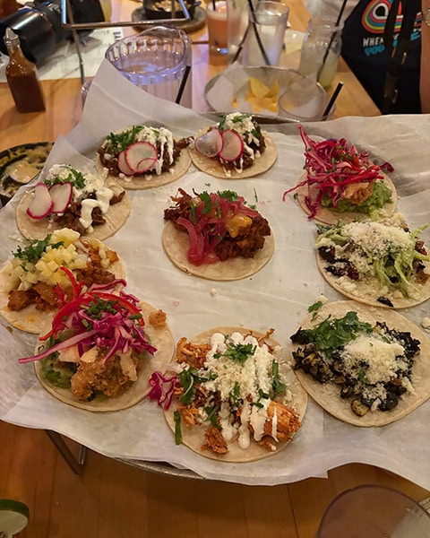Eine Auswahl von neun verschiedenen mexikanischen Tacos, die kreisförmig auf einem großen, mit weißem Papier ausgelegten Servierteller in einem Restaurant angerichtet sind. Die Tacos zeigen vielfältige Beläge, darunter gezupftes Fleisch, leuchtend rosa eingelegte rote Zwiebeln, frische Radieschenscheiben, Kräuter wie Koriander und fein geriebenen weißen Käse. Im Hintergrund sind Restauranttische mit Getränkegläsern und einer Flasche Chilisauce unscharf zu sehen.