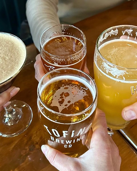 Vier Personen stoßen an einem Holztisch mit verschiedenen Kaltgetränken an. Im Vordergrund ist ein Bierglas mit der Aufschrift „Tofino Brewing Co.“ zu sehen, daneben zwei weitere Biergläser und ein Cocktailglas.