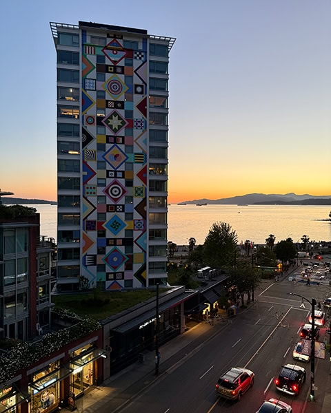 Blick von oben auf eine städtische Straße bei Sonnenuntergang in Vancouver. Links steht ein markantes Hochhaus mit einer bunten, geometrisch gemusterten Fassade. Im Hintergrund ist das Meer der English Bay mit Silhouetten von Bergen am Horizont zu sehen.