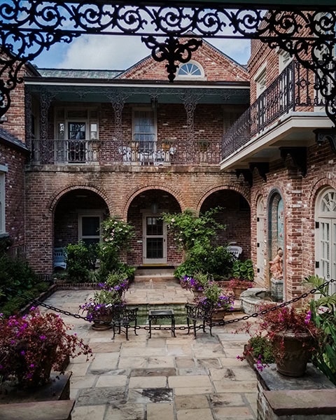 Ein gepflasterter, mit Blumenkübeln und Gartenmöbeln gestalteter Innenhof im französischen Stil liegt vor der historischen, zweistöckigen Backsteinfassade des Bellingrath Home, geschmückt mit kunstvollen, gusseisernen Balkongeländern im Vordergrund.