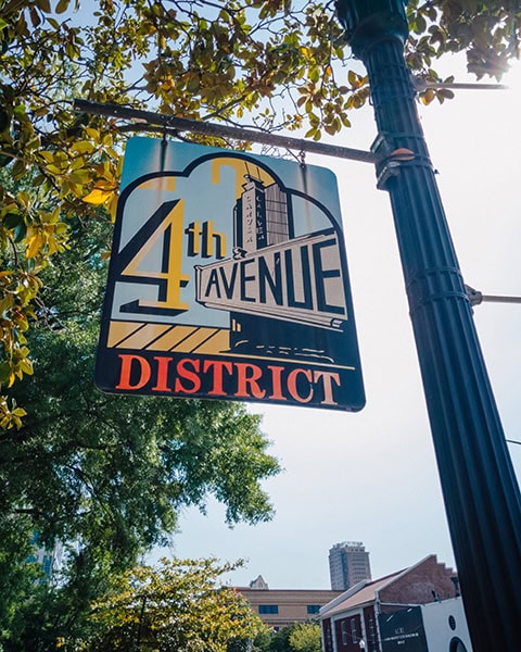 Ein kunstvoll gestaltetes Schild mit der Aufschrift „4th AVENUE DISTRICT“ hängt an einem gusseisernen Pfosten vor grünen Baumkronen, während im fernen Hintergrund historische Gebäude unter blauem Himmel aufragen.