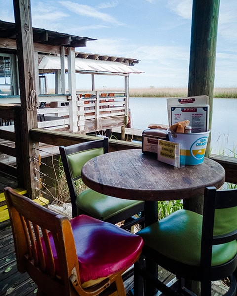 Ein runder Holztisch mit bunten Lederstühlen steht auf der überdachten Außenterrasse des BLUEGILL Restaurant in Spanish Fort, bestückt mit Speisekarten in einem Eimer, während man im Hintergrund direkt auf das ruhige Gewässer des Mobile-Tensaw River Delta blickt.