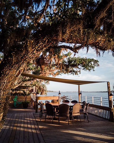 Ein runder Esstisch mit geflochtenen Stühlen steht auf der hölzernen, im warmen Abendlicht liegenden Außenterrasse des beliebten Küstenrestaurants Cap's on the Water in St. Augustine, Florida, überspannt von einem beigen Sonnensegel und den massiven, mit Lichterketten verzierten Ästen einer alten Eiche, während im Hintergrund der ruhige Intracoastal Waterway (Tolomato River) verläuft.
