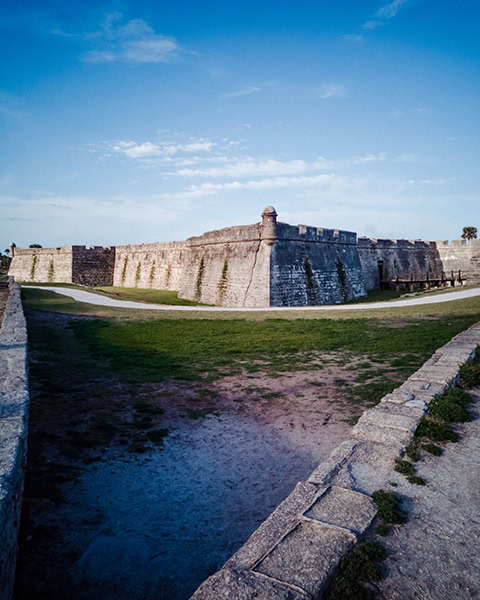 Die massiven, grauen Steinmauern der historischen Festung Castillo de San Marcos in St. Augustine, Florida, ragen mit einer charakteristischen Ecktrompete (Scharwachturm) hinter einem breiten Befestigungsgraben auf, während sich im Vordergrund eine alte Steinmauer und ein geschwungener Fußweg unter einem blauen Himmel erstrecken.