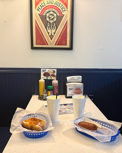Ein mit Chili und Käse belegter Hotdog und ein Corn Dog liegen jeweils in blauen Plastikkörbchen auf einem hellen Restauranttisch, auf dem zwei große weiße Becher sowie Saucenflaschen stehen, während an der Wand darüber ein gerahmtes Plakat mit der Aufschrift „PEACE AND JUSTICE“ hängt.