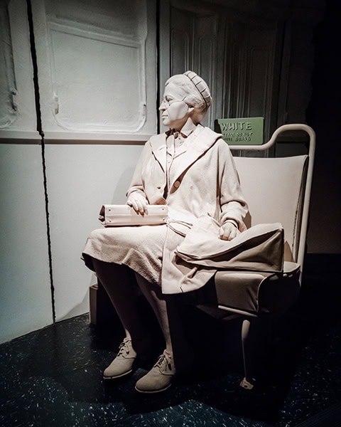 Eine lebensgroße, einfarbig helle Figurennachbildung von Rosa Parks sitzt mit Brille, Mantel und einer Handtasche auf einem Bussitz, während sie aus dem angedeuteten Fenster eines historischen Linienbusses blickt.