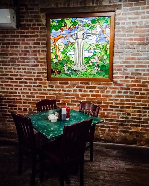 Ein quadratischer Holztisch mit vier dunklen Stühlen steht vor einer rustikalen, roten Backsteinwand im legendären Clary's Cafe in Savannah, Georgia. An der Wand hängt ein von innen beleuchtetes, farbenfrohes Buntglasfenster, das die berühmte „Bird Girl“-Statue inmitten von Bäumen und rosa Blüten zeigt, während auf der grünen, marmorierten Tischplatte Menagerien mit Saucen bereitstehen.