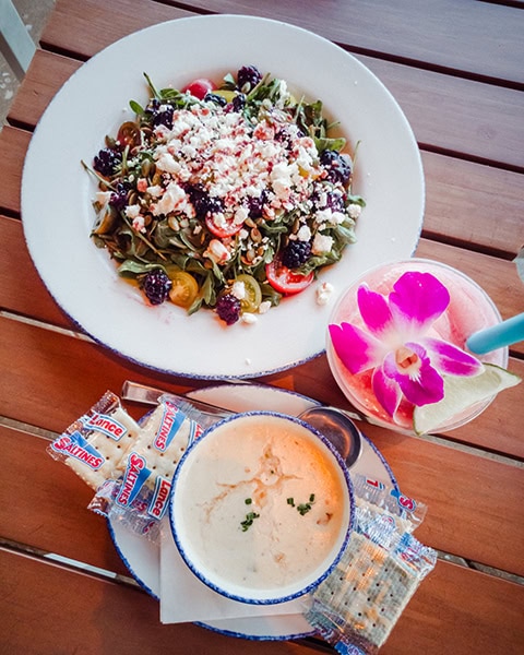 Ein großer, frischer Salat mit Rucola, Brombeeren, Kirschtomaten und Feta-Käse, eine cremige Suppe mit Lance Saltines-Crackern und ein mit einer lila Orchidee garnierter Cocktail stehen servierfertig auf einem Holztisch im Restaurant The CoastAL.