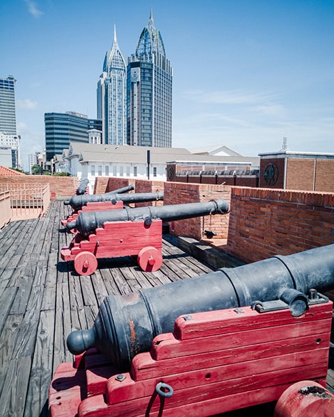 Eine Reihe historischer Kanonen auf roten Holzlafetten steht auf der hölzernen Aussichtsplattform der Festungsrekonstruktion des Fort Condé, während im Hintergrund moderne Wolkenkratzer wie der RSA Battle House Tower in Mobile emporragen.