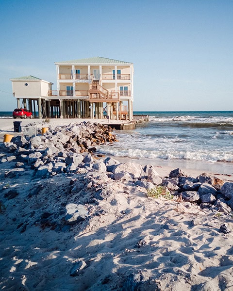 Ein mehrstöckiges Strandhaus auf hohen Holzpfählen steht direkt am Ufer des Golfs von Mexiko in Dauphin Island, geschützt von einer steinernen Ufermauer und Sanddünen im Vordergrund unter einem klaren blauen Himmel.
