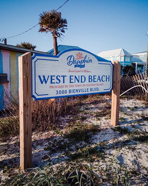 Ein großes Holzschild am sandigen Strandzugang weist mit blau-weißer Schrift auf den West End Beach in Dauphin Island hin, umgeben von Strandgräsern, einer Palme und bunten Küstenhäusern im Hintergrund unter klarem blauem Himmel.