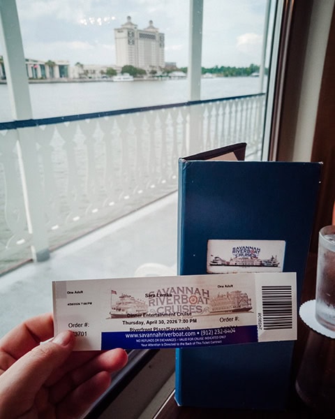 Eine Hand hält ein Papierticket für eine „Dinner Entertainment Cruise“ von Savannah Riverboat Cruises im Vordergrund vor einer blauen Speisekarte, während man durch das Fenster des Raddampfers über das weiße Geländer hinweg direkt auf den Savannah River und das markante Hochhaus des The Westin Savannah Harbor Golf Resort & Spa am gegenüberliegenden Ufer blickt.