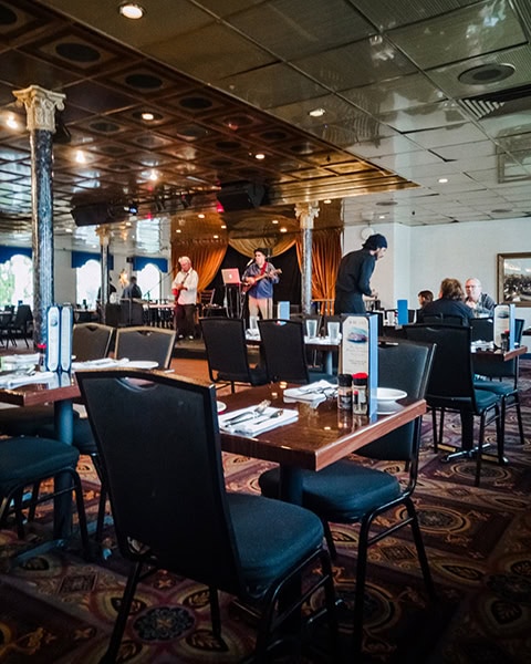 Der weitläufige, elegant eingerichtete Speisesaal im Inneren des Raddampfers „Georgia Queen“ von Savannah Riverboat Cruises zeigt eingedeckte Holztische mit dunklen Stühlen auf einem gemusterten Teppichboden unter einer glänzenden Kassettendecke, während im Hintergrund eine Live-Band mit Gitarristen auf einer kleinen Bühne mit goldenen Vorhängen für die Abendunterhaltung der Gäste spielt.