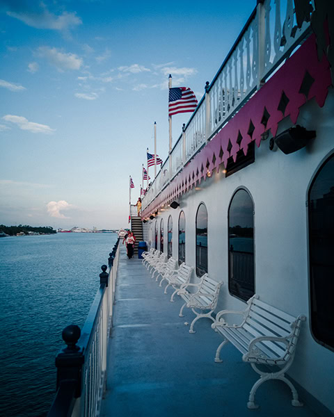 Das lange Außendeck des historischen Raddampfers „Georgia Queen“ von Savannah Riverboat Cruises erstreckt sich entlang des Savannah River im Abendlicht, bestückt mit einer Reihe weißer, kunstvoller Parkbänke vor großen Bogenfenstern. Am oberen, weiß-rot verzierten Relingsbereich wehen mehrere US-amerikanische Flaggen im Wind, während in der Ferne Passagiere auf dem Deck spazieren gehen.