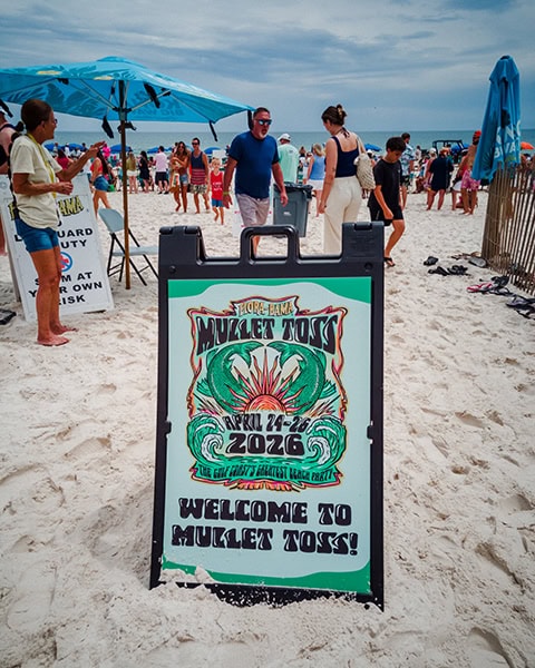 Ein großer, schwarzer Aufsteller steht im weißen Sandstrand am Flora-Bama Lounge & Oyster Bar Strandabschnitt und zeigt ein buntes Plakat mit der Aufschrift „Flora-Bama Mullet Toss 2026“, während im Hintergrund zahlreiche Strandbesucher und Sonnenschirme vor dem Meer unter bewölktem Himmel zu sehen sind.