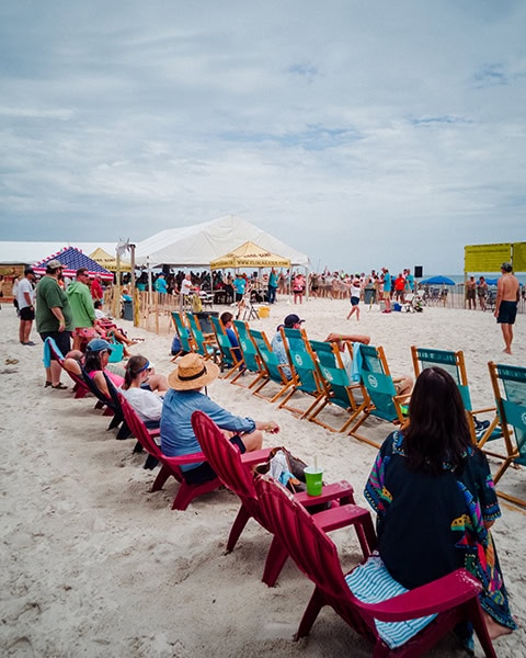 Zahlreiche Zuschauer sitzen in einer langen Reihe von roten und türkisfarbenen Strandstühlen im weißen Sand und verfolgen das Geschehen beim jährlichen „Interstate Mullet Toss“-Wettbewerb am Strand der Flora-Bama Lounge, während im Hintergrund ein großes weißes Veranstaltungszelt und eine Menschenmenge unter wolkenverhangenem Himmel zu sehen sind.