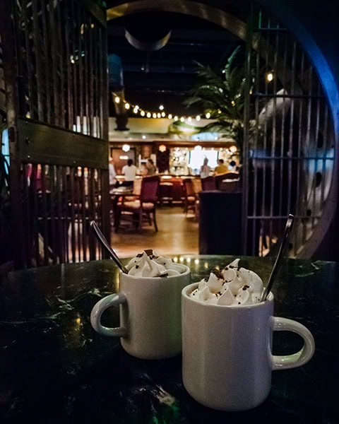 Zwei weiße Kaffeetassen mit reichlich Schlagsahne, Kakaopulver und Schokoladenstückchen stehen im Vordergrund auf einem dunklen Marmortisch, während im unscharfen Hintergrund ein beleuchteter Barbereich hinter einer geöffneten, runden Tresortür zu sehen ist.