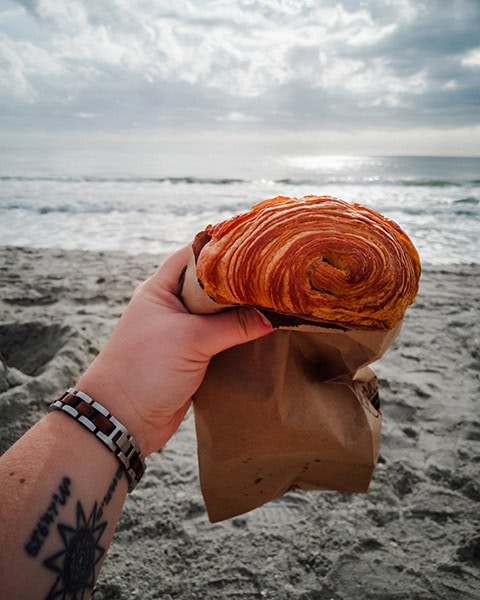 Eine Hand mit einem tätowierten Handgelenk hält ein großes, goldbraun gebackenes und perfekt geschichtetes Croissant (Pain au Chocolat) in einer braunen Papiertüte im Vordergrund, während im Hintergrund die Wellen des Atlantischen Ozeans an den Sandstrand von Tybee Island (nahe Savannah, Georgia) schlagen.