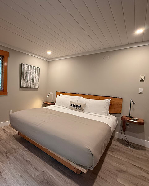 Ein modernes Hotelzimmer mit hellem Holzboden und einer weiß getäfelten Decke. Im Zentrum steht ein großes Bett mit einer beige-weißen Decke und einem Kopfteil aus einer massiven Baumkante. An den Seiten hängen Nachttische aus Naturholz mit schwarzen Lampen, links an der Wand hängt ein quadratisches Bild mit Birkenstämmen.