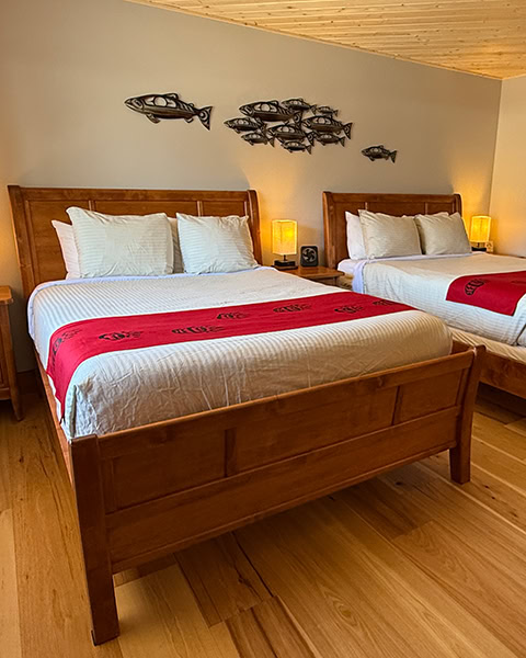 Ein gemütliches Hotelzimmer mit hellem Holzboden und einer hellen Holzdecke. Im Raum stehen zwei große Betten aus warmem Holz mit weißer Bettwäsche und roten Bettläufern, die mit Mustern im First-Nations-Stil verziert sind. An der Wand über den Betten hängt eine dekorative Wandkunst aus Metall, die einen Schwarm Lachse darstellt.