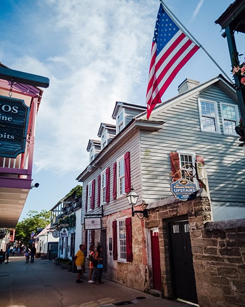 Eine große Flagge der Vereinigten Staaten hängt an der Fassade des historischen Rodriguez-Avero-Sanchez House in der bekannten St. George Street in St. Augustine, Florida, während sich im Erdgeschoss das steinerne Mauerwerk zeigt und Passanten entlang der historischen Fußgängerzone schlendern.